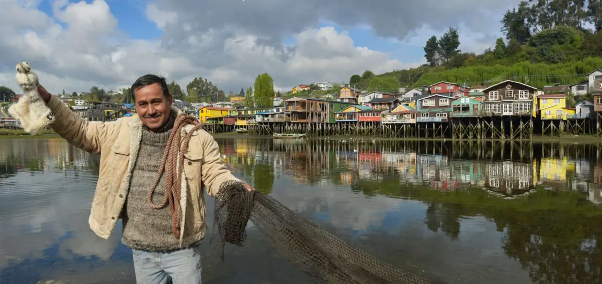 Hombre con redes frente a los palafitos de colores de Chiloé reflejados en el agua