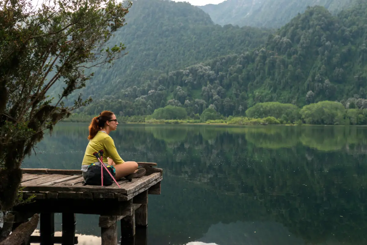 Mujer sentada en un muelle de madera mirando el lago Ranco, rodeado de cerros cubiertos de bosque