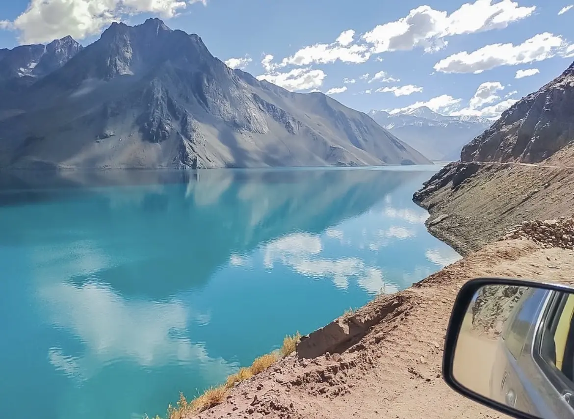 Lago de aguas turquesas entre montañas, visto desde la ventana de un auto en un camino de la cordillera