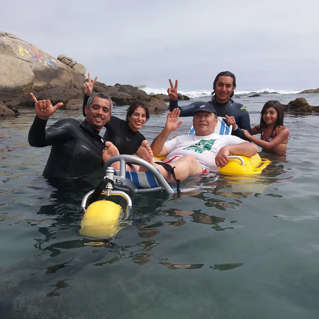 Personas con silla anfibia y acompañantes bañándose en el mar en la playa inclusiva Las Conchitas
